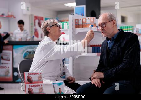 Apothecary worker holding infrared thermometer near elderly customer forehead, testing patient for fever. Old customer measuring body temperature in drugstore, getting common cold diagnosis Stock Photo