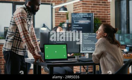 Programer working at desk with multiple computer screens and laptop with green screen chroma key mockup running code. Software developer compiling source algorithm interrupted by colleague. Stock Photo