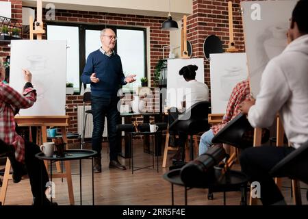Senior man experienced artist teaching drawing classes to adults, creating positive learning environment in art class. Group of diverse different aged students practice sketching skills together Stock Photo