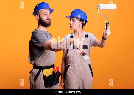 Young construction workers posing with painting tools, standing over yellow background. Team of constructors holding paintbrush and roller brush to work on refurbishment project in studio. Stock Photo