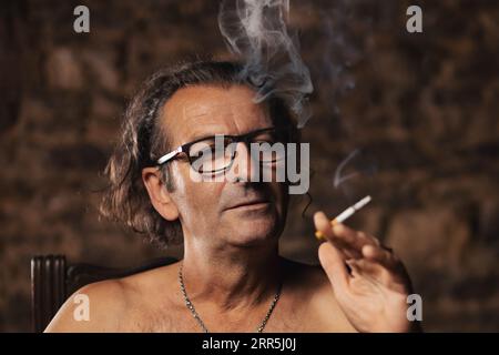 An older man, about 60 years old, of Serbian nationality with long hair and glasses, thoughtfully smokes a cigarette on a dark background. Artist, cre Stock Photo