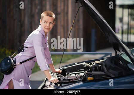 A woman near a broken car in front of the open hood looks to the side Stock Photo
