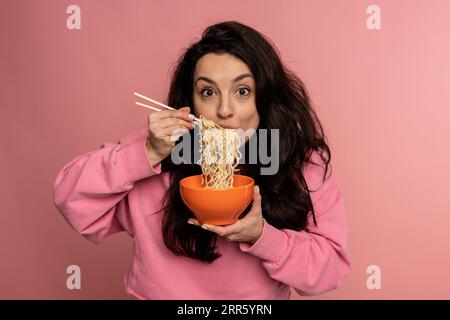 Hungry dark-haired young Caucasian woman eating pasta with an appetite during the studio photo shoot on the pink background. Japanese food concept Stock Photo