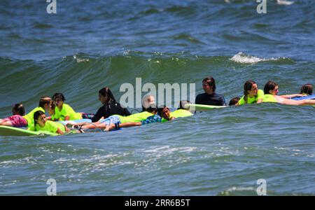 Gilgo Beach, New York, USA - 25 July 2023: Kids in neon green shirts trying to ride on waves with their counselors help while surfing at Bunger Surf C Stock Photo