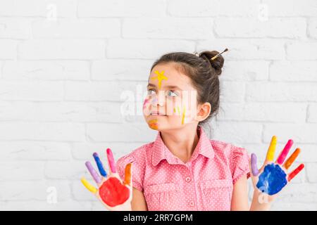 Portrait girl standing against white wall showing colorful painted hands looking away Stock Photo
