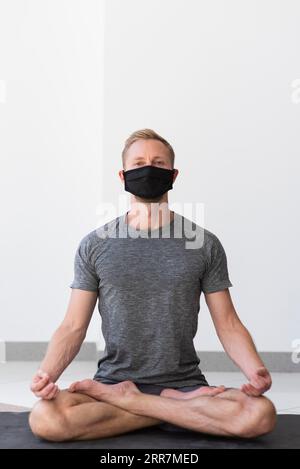 Full shot man with face mask doing sukhasana pose inside mat Stock Photo