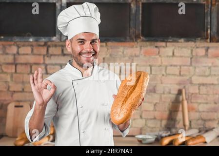 Smiling male baker holding loaf showing ok hand sign gesture Stock Photo