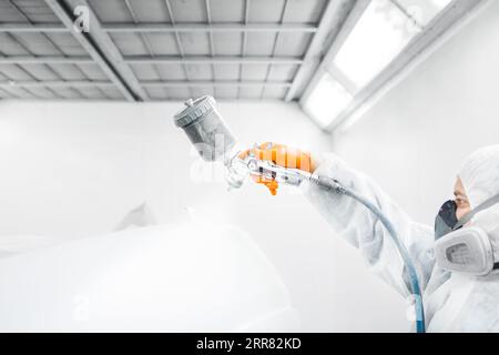 Male worker in protective clothes and mask painting car using spray paint. Stock Photo