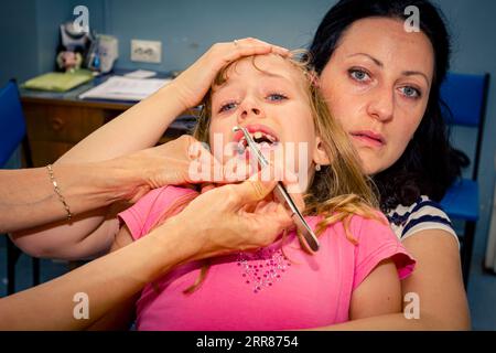 Preschooler, scared and crying cute little child is sitting on mom's lap at dentist office, doctor holds tweezers, prepare for removing milk tooth. Stock Photo