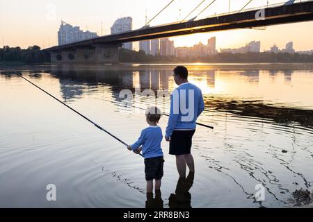 Father and son are fishing together standing with fishing rods in the water of the river at sunset. father's day concept. Family fun pastime for the w Stock Photo