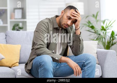 Upset african american man sitting on sofa at home and holding his head. Thoughtfully looks to the side, suffers from depression. Stock Photo