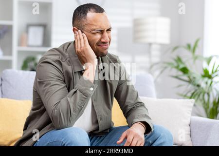 Severe pain and inflammation in the ear of a young African American man sitting on the couch at home and holding his head. Stock Photo