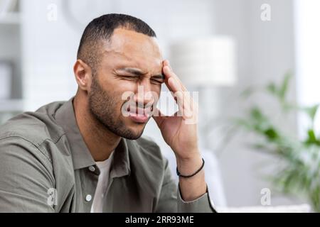 Close-up photo of a young African American man suffering from a severe headache. Sitting at home on the sofa, grimacing in pain, holding his head with his hand, doing a massage. Stock Photo