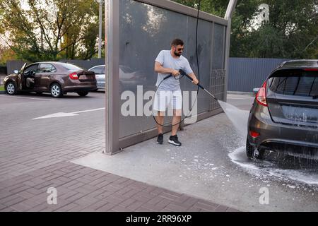 At the car wash, a man washes the foam foam off the bottom of the car with a water cannon.  A car at a self service car wash. Stock Photo