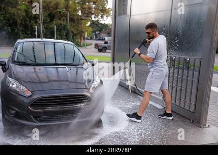 The man aims the water cannon at the car's front headlights and washes them. A car at a self service car wash. Stock Photo