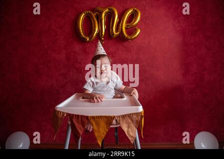 Portrait of Little boy of one year in festive hat sits in childrens chair with fabric flags. Kid on red isolated background with foil gold balloons On Stock Photo