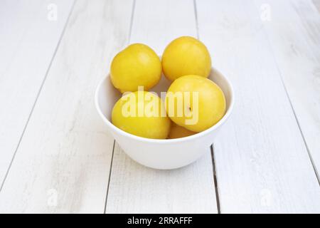 A small white porcelain bowl filled with ripe yellow plums on a white wooden table Stock Photo