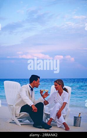 Maldives. Young couple in evening clothes sitting, drinking Champagne on the beach at dusk. Stock Photo