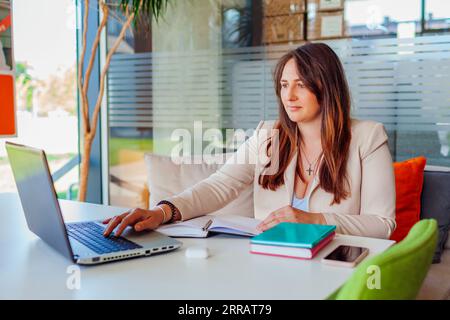 Portrait of young businesswoman working in office using laptop. Middle-aged woman writing down notes in notebook Stock Photo