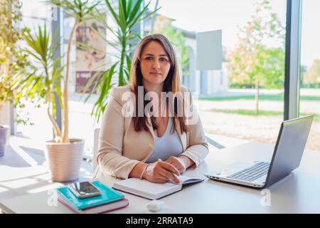 Happy businesswoman working online in office using laptop. Smiling middle-aged woman looking at camera sitting at desk typing. Light workplace with gl Stock Photo
