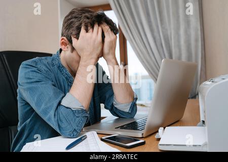 Tired stressed young man sitting in front of laptop, frustrated with problems or study failure, feel exhausted, having headache, upset with bad news, money problem, unsuccessful exam or test results. Stock Photo