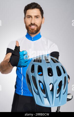 Smiling bearded cyclist in activewear showing thumb up to camera. Front view of male biker demonstrating blue cycle helmet, approving, isolated on white studio background. Concept of cycling activity. Stock Photo
