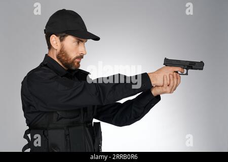 Focused bearded police officer taking aim, pointing with handgun in studio. Side view of caucasian cop shooting, holding gun, looking away, isolated on gray background. Concept of danger work, weapon. Stock Photo