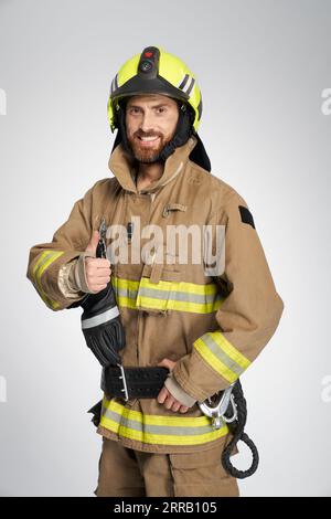 Professional firefighter showing thumb up to camera in studio. Portrait of handsome bearded fireman in uniform gesturing, giving like and smiling, on gray background. Concept of work, hand gesture. Stock Photo