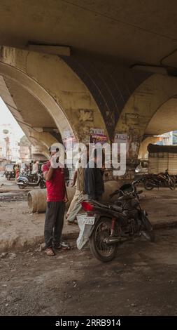 Young Indian male calling with his mobile phone standing next to his motorbike, under a bridge, while three elderly men walk by in Aurangabad, India. Stock Photo