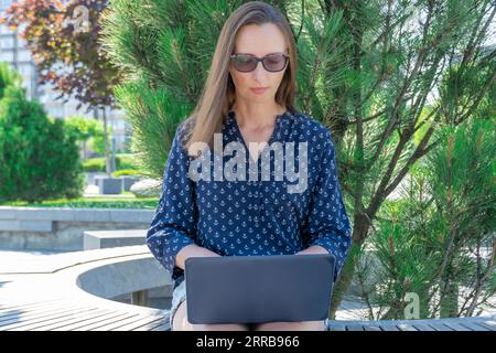 Woman freelance works on laptop in city park. Young adult student education in fresh air. Female sits outdoor and types on computer. Workplace on natu Stock Photo
