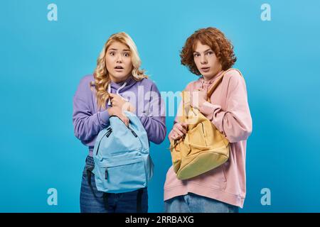 scared and worried teenage students in hoodies looking at camera and standing with backpacks on blue Stock Photo