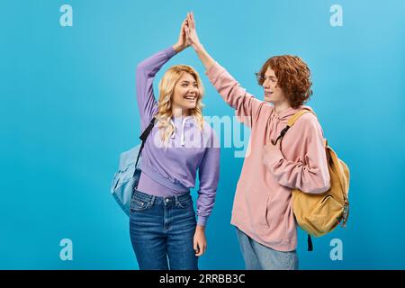 happy teenage classmates with backpacks wearing trendy clothes and giving high five on blue Stock Photo