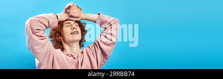 dreamy, smiling redhead teen boy in hoodie with closed eyes and hands on forehead on blue, banner Stock Photo