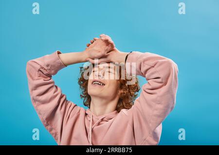 positive, dreamy redhead teen guy in hoodie standing with closed eyes and hands on forehead on blue Stock Photo