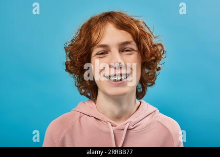 carefree and joyful teenage guy with red wavy hair and braces looking at camera on blue, portrait Stock Photo