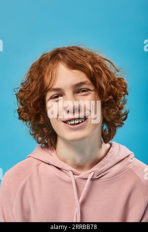 portrait of cheerful teenage guy with red wavy hair and braces smiling at camera on blue, happiness Stock Photo