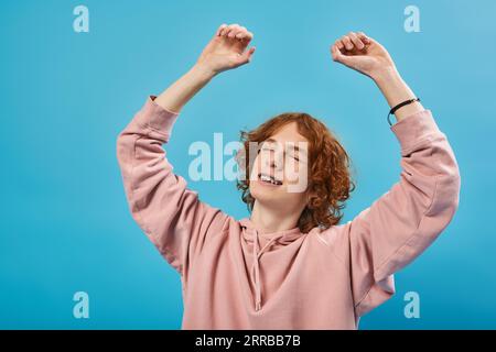 smiling and dreamy redhead teen guy in hoodie standing with closed eyes and raised hands on blue Stock Photo