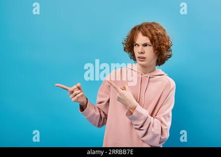 displeased redhead teenage boy in trendy hoodie looking away and pointing with fingers on blue Stock Photo