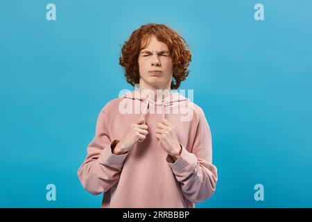 nervous boy with red wavy hair wearing trendy hoodie and standing with closed eyes on blue Stock Photo