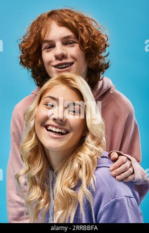 portrait of carefree teenage friends, redhead guy and blonde girlfriend looking at camera on blue Stock Photo