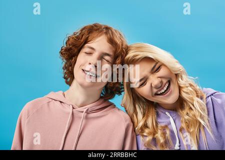 portrait of excited teenage boy and girl in hoodies laughing with closed eyes on blue, happiness Stock Photo