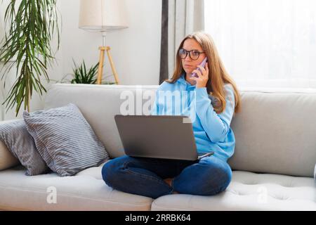 Woman freelancer in glasses using phone and laptop while sitting on couch Stock Photo