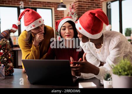 Women workers team gathered around laptop, discussing project tasks in office during christmas holiday. Diverse colleagues in santa hats collaborating on annual report on xmas eve Stock Photo