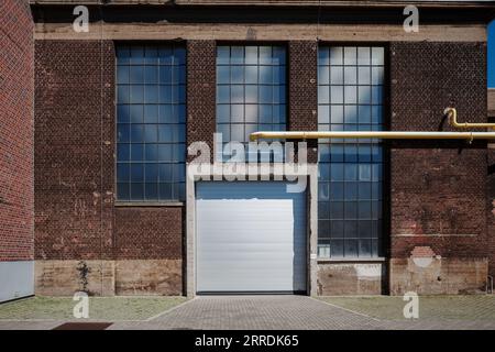 Outdoor exterior street view in front of historical industrial building with red old rough brick facade, tall glass windows and white metal shutter. Stock Photo