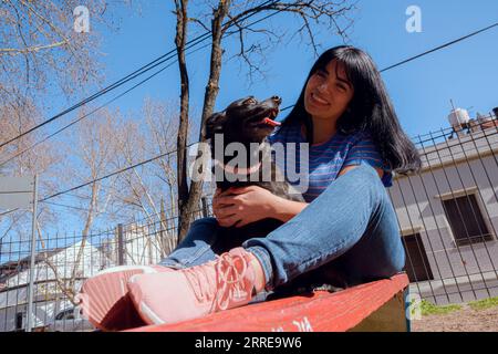 happy young black haired latin woman sitting on bench with her dog smiling and playing in park at sunset, enjoying day, copy space. Stock Photo