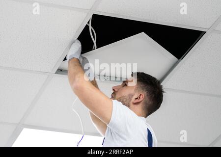 Full Length Portrait Of Electrician On Stepladder Installs Lighting To The Ceiling In Office Stock Photo