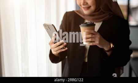 Cropped image of a happy and smiling Asian Muslim businesswoman in a formal black suit and hijab is checking messages on her smartphone and having cof Stock Photo