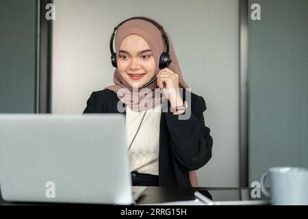 A friendly and beautiful Asian Muslim female call centre operator or technical support wearing a headset and hijab is answering an incoming call and w Stock Photo