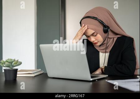 A stressed and tired Asian Muslim female call centre operator or customer support representative wearing a headset and hijab is worried about a custom Stock Photo
