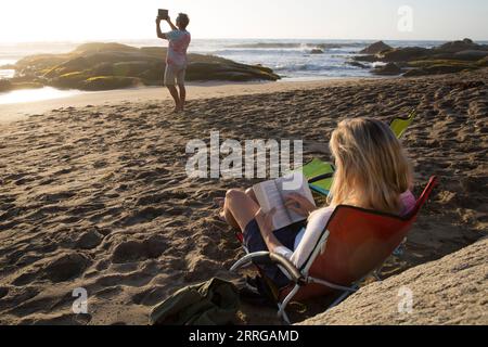 Mature woman reads book in beach chair as man takes photo with tablet Stock Photo